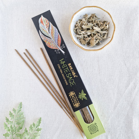 Caja de incienso de Salvia Blanca de la marca Banjara con la figura del pájaro del Trueno. A su lado, varias varillas de incienso sueltas sobre una superficie de piedra clara, con una de ellas encendida y un suave humo elevándose.