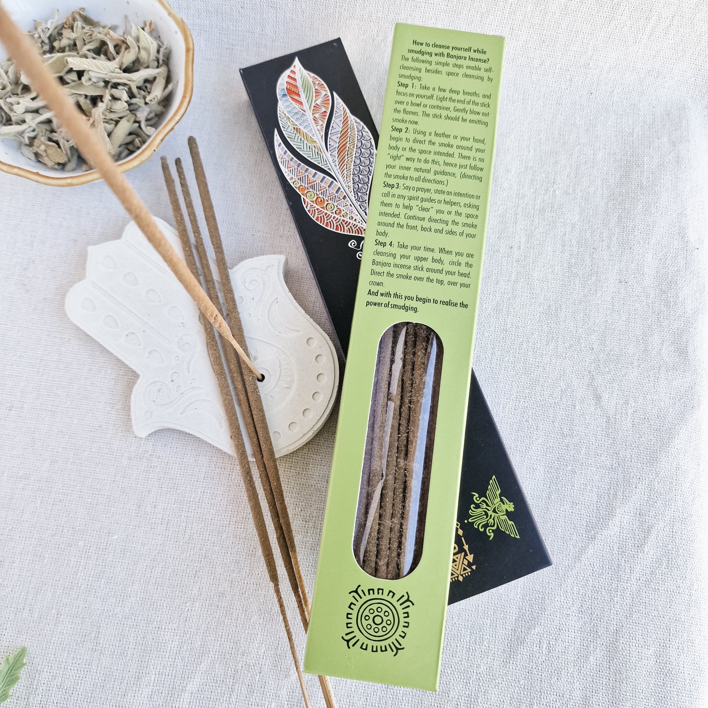 Caja de incienso de Salvia Blanca de la marca Banjara con la figura del pájaro del Trueno. A su lado, varias varillas de incienso sueltas sobre una superficie de piedra clara, con una de ellas encendida y un suave humo elevándose.