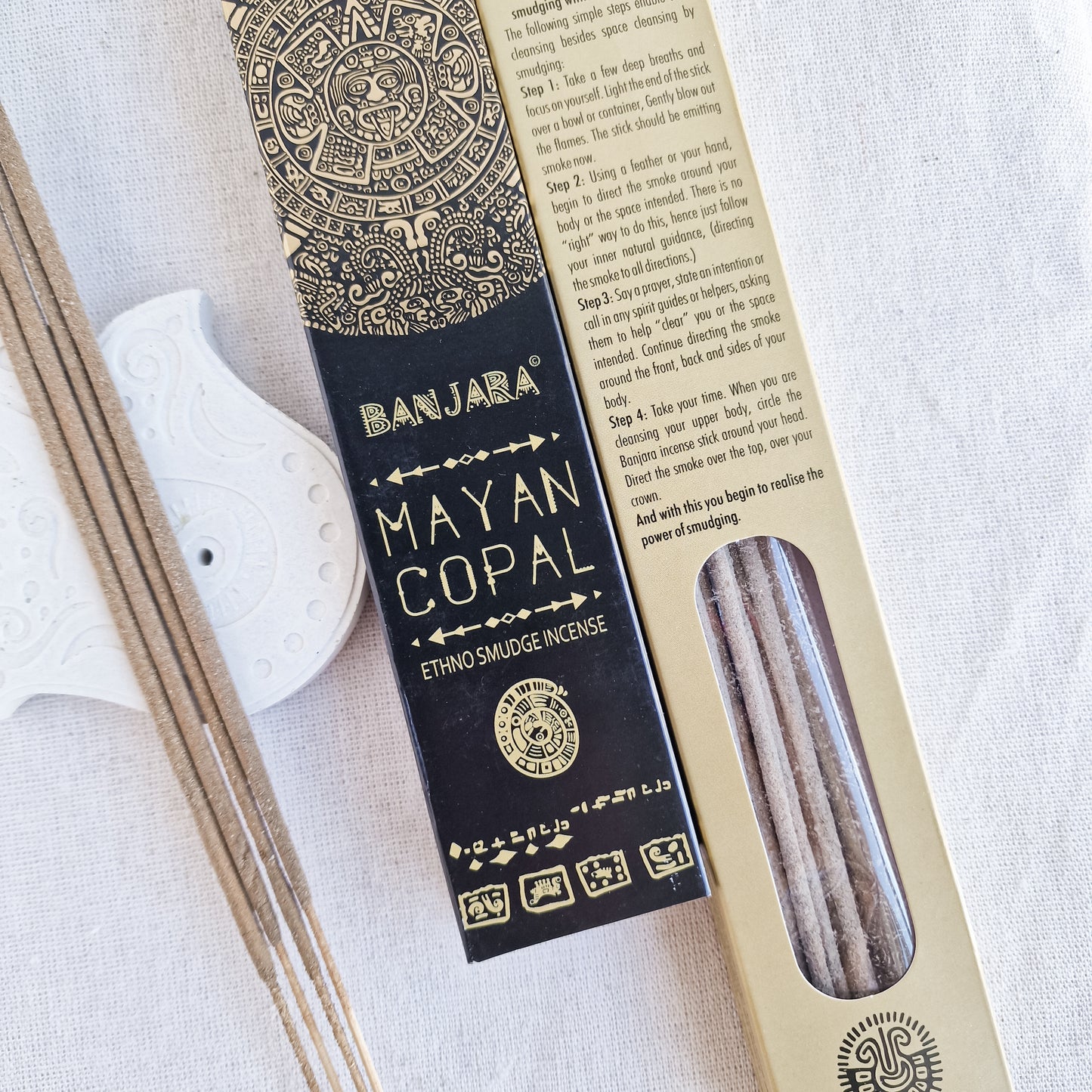 Caja de incienso de Copal de la marca Banjara con la figura de la serpiente emplumada Quetzalcóatl. A su lado, varias varillas de incienso sueltas sobre una superficie de piedra clara, con una de ellas encendida y un suave humo elevándose.