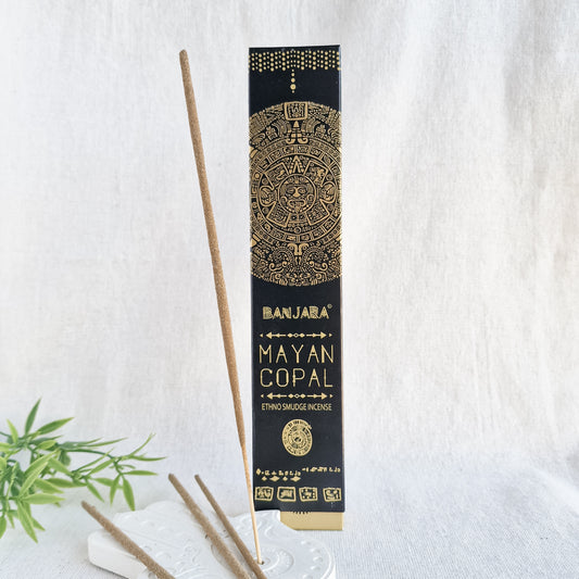 Caja de incienso de Copal de la marca Banjara con la figura de la serpiente emplumada Quetzalcóatl. A su lado, varias varillas de incienso sueltas sobre una superficie de piedra clara, con una de ellas encendida y un suave humo elevándose.