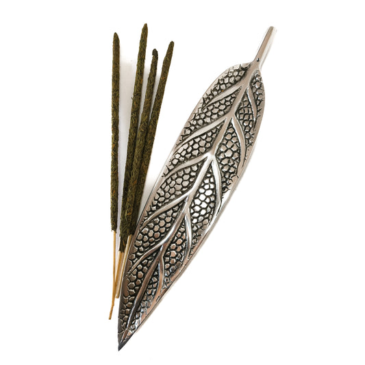 Un porta incienso de metal con forma de hoja, diseñado para sostener una varilla y recoger la ceniza. La varilla de incienso está encendida y el humo se eleva suavemente.