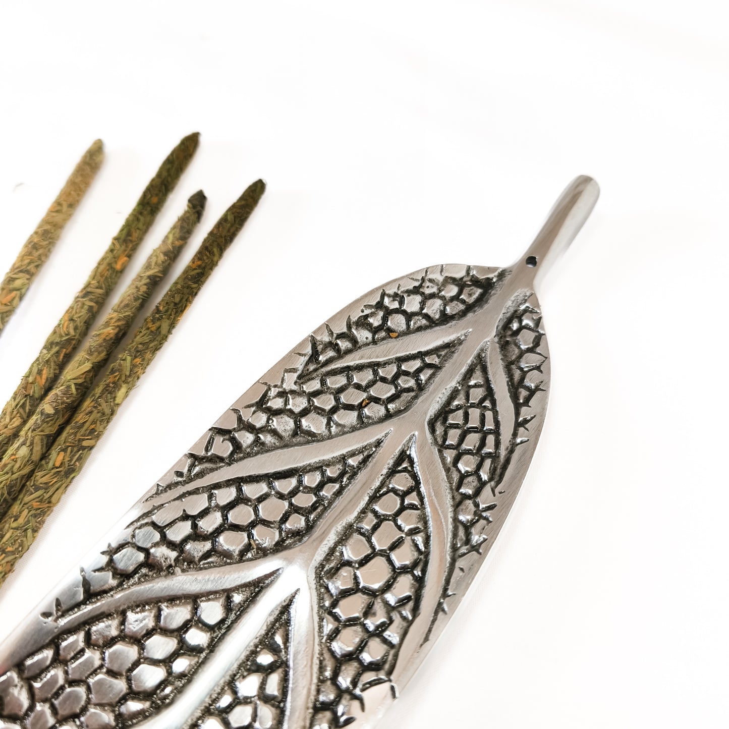 Un porta incienso de metal con forma de hoja, diseñado para sostener una varilla y recoger la ceniza. La varilla de incienso está encendida y el humo se eleva suavemente.