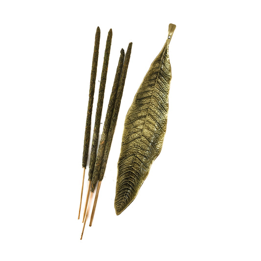 Un porta incienso de metal pequeño con forma de hoja en color bronce. La varilla de incienso está encendida sobre la hoja.